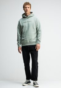 Lichtgroene hoodie met de tekst "BUTCHER OF BLUE", zwarte jeans en zwart-witte Converse sneakers, met een relaxte pasvorm en een trekkoord in de capuchon.