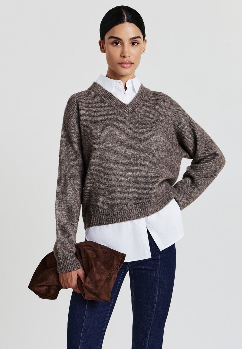 Maglione marrone a trama con scollo a v ampio, abbinato a una camicia bianca con colletto. Il modello tiene in mano una pochette in suede marrone. Jeans scuri completano l'outfit.