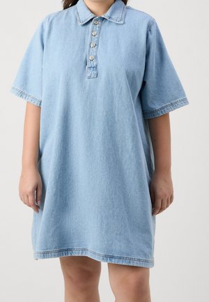 Kobieta ubrana w luźną, jasnoniebieską sukienkę z denimu z krótkimi rękawami, kołnierzykiem i czterema guzikami na przodzie.