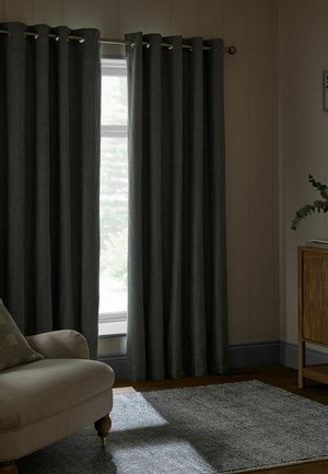 Dark gray curtains partially open on a window with soft natural light, beige armchair, gray rug, and wooden cabinet with a plant in a living room.