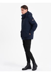 Námořnická modrá zateplená parka s kapucí, se zapínáním na knoflíky vpředu, dvěma velkými kapsami a logem na rukávu.