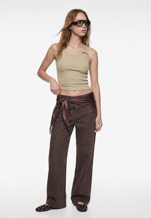 Mujer con top asimétrico beige, pantalones marrones de pierna ancha con un pañuelo de encaje morado oscuro anudado, gafas de sol y zapatos planos negros con tachuelas.