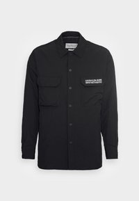 Black long-sleeved shirt featuring a button-up front, two chest pockets, and "Calvin Klein Jeans" embroidered in white on the right pocket.