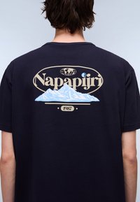 Navy blue cotton t-shirt featuring a mountain graphic and "Napapijri" text in beige, with a round logo and the year "1987" underneath.