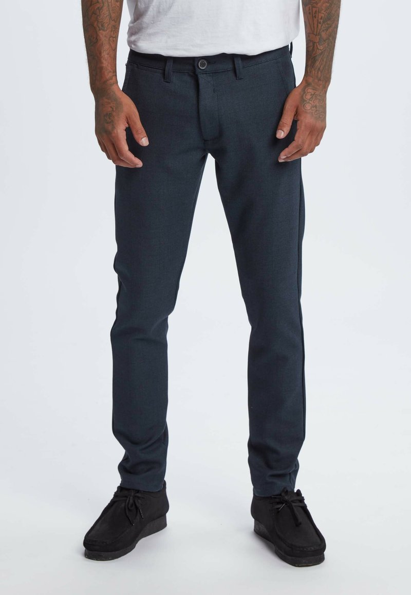 Dark blue trousers made of textured fabric featuring a slim fit, belt loops, and a single button closure. Worn with black shoes.