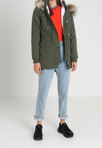 Parka verde oliva con capucha con pelo, que cuenta con un cierre de cremallera y botón, bolsillos laterales y un forro interior de forro polar. Usada con unos vaqueros celestes remangados y zapatillas negras.