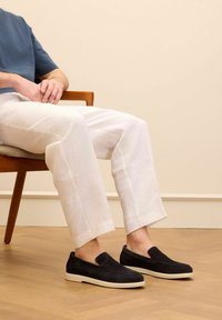 Black suede slip-on shoes with a white sole, paired with white linen pants. The shoes feature a smooth, sleek design and no visible hardware.