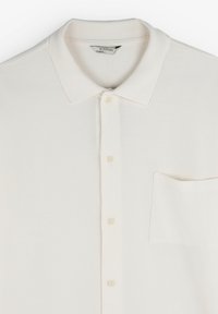 Cămașă polo de culoare crem, realizată dintr-un material texturat, având un guler clasic, cinci butoane și un singur buzunar tip patch.