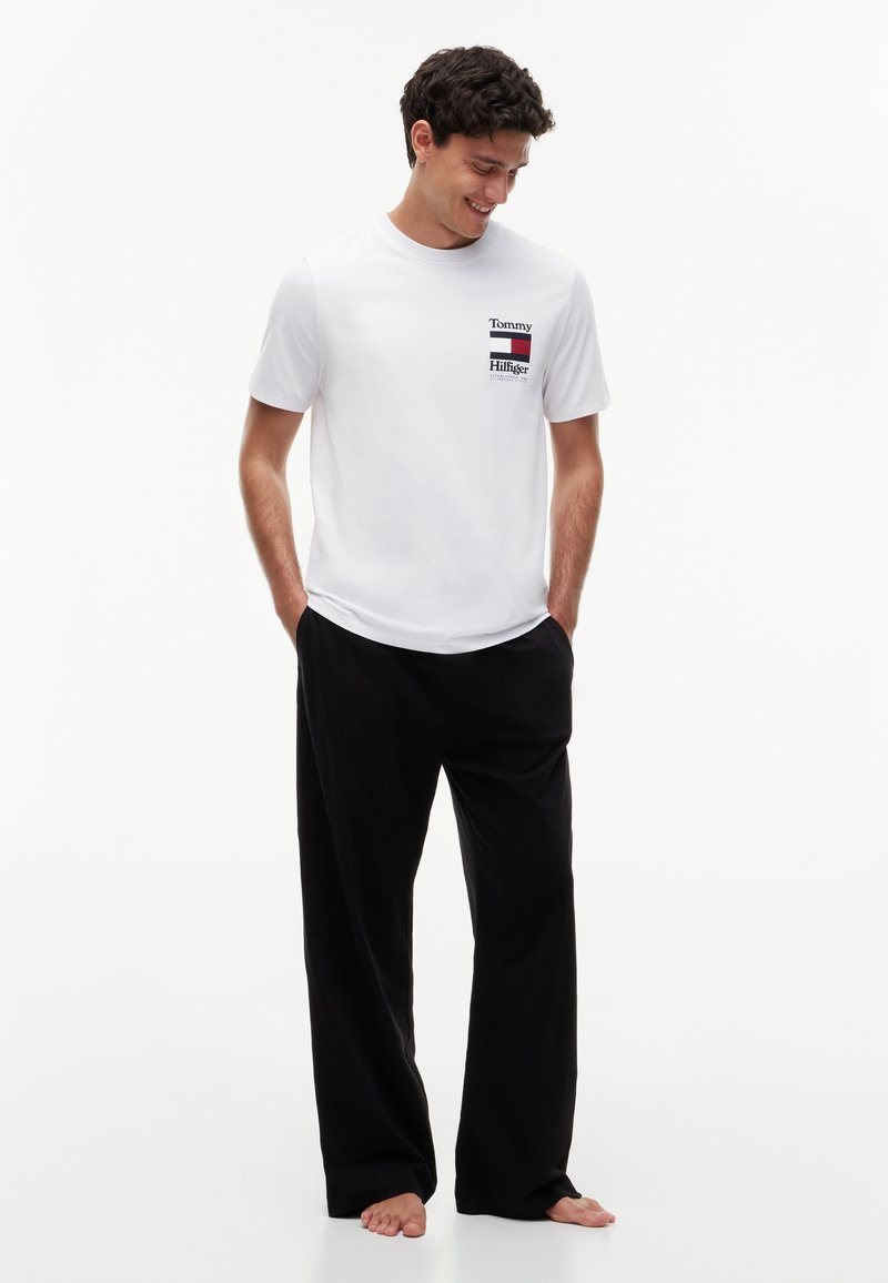 Jeune homme debout pieds nus portant un t-shirt blanc Tommy Hilfiger et un pantalon noir ample, les mains dans les poches, regardant vers le bas avec un sourire.