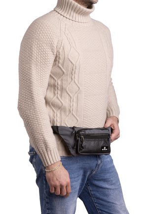 Maglione beige lavorato a maglia con collo alto. Borsa a cintura nera con molte tasche con zip indossata in vita sopra un paio di jeans blu.