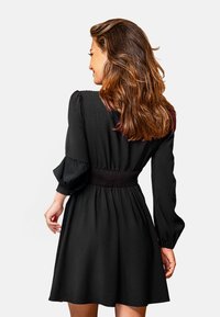 Vestido negro con cintura smock, mangas largas abullonadas y falda acampanada. Tejido suave y diseño sencillo. Sin patrones o acentos visibles.