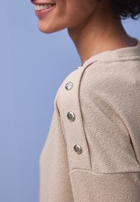 Personne souriante portant un pull en maille beige avec des boutons décoratifs en argent le long de l'épaule, sur un fond bleu.