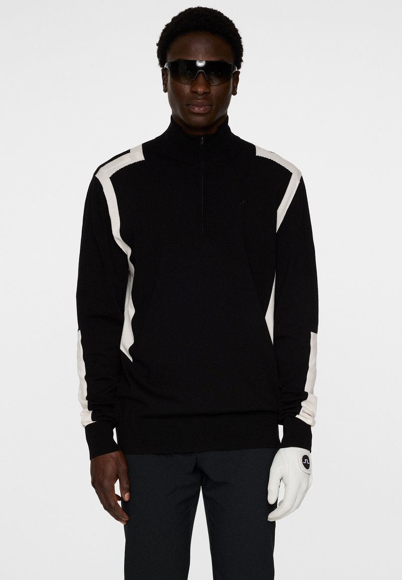 Svart kvartdraget pullover med vita diagonala detaljer på ärmarna. Har en mjuk textur och ribbad krage. Modellen bär vita handskar och solglasögon.