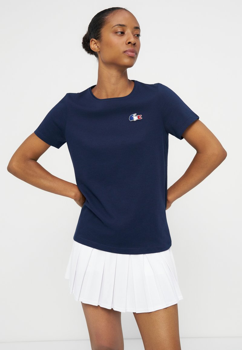 Jeune femme portant un T-shirt bleu marine avec un petit logo et une jupe plissée blanche, debout les mains sur les hanches devant un fond uni.