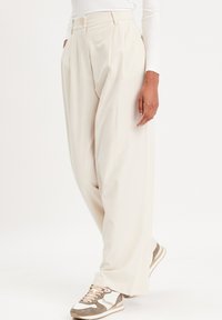 Pantalons beige à jambe large avec taille haute, présentant des plis à l'avant et une fermeture à un bouton. Assortis avec des baskets beige et gris.
