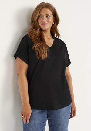 Vrouw met lang rood haar, gekleed in een zwart kort mouw V-hals topje en blauwe jeans, staand tegen een lichte muur, kijkt opzij.
