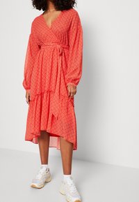 Punainen kuvioitu midi-mekko, jossa on kietaisusuunnittelu, pitkät puffihihat ja kerroksellinen helma. Yhdistetty valkoisiin ja beigeihin paksupohjaisiin lenkkarikenkiin.