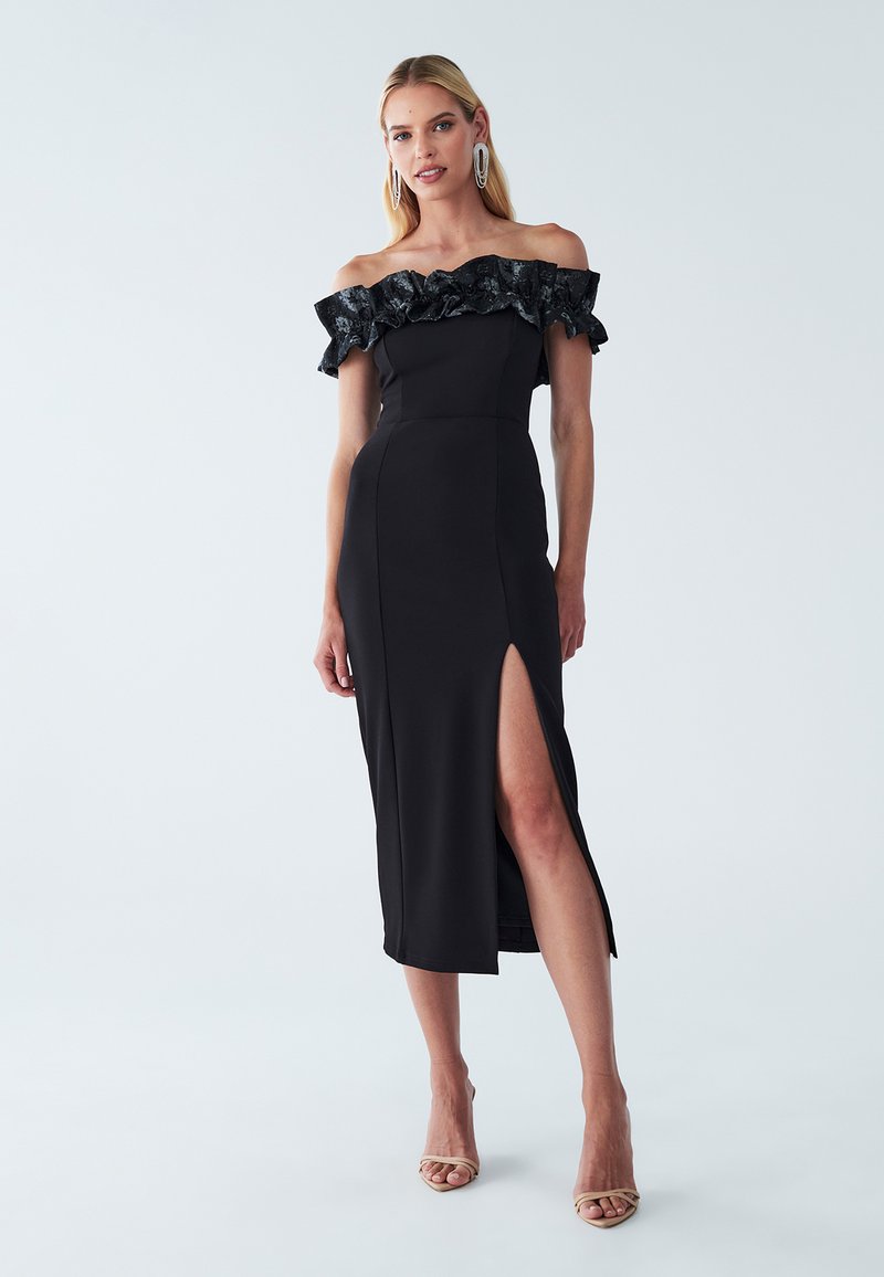 Black off-the-shoulder dress with ruffled edges, thigh-high slit, body-hugging fit, and smooth texture; worn with strappy beige heels.