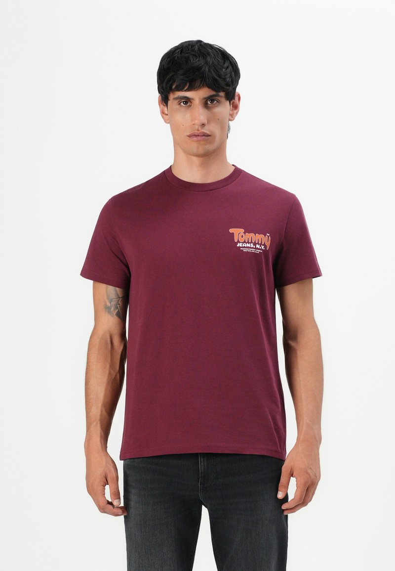 Mann in einem schlichten burgunderfarbenen T-Shirt mit dem Logo "Tommy Jeans, N.Y." auf der linken Brust, kombiniert mit dunklen Jeans und stehend vor einem weißen Hintergrund.