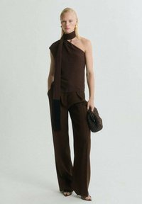 Top marrone senza maniche con dettaglio di sciarpa, abbinato a pantaloni marroni a gamba larga. La modella tiene in mano una borsa marrone con texture e indossa scarpe aperte.