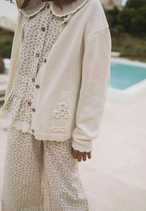 Niño con cárdigan de punto color crema con bordado floral sobre blusa beige con flores y pantalones a juego, cerca de una piscina al aire libre.