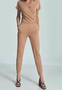 Combinaison couleur beige avec manches courtes, devant croisé, taille élastique et poches latérales ; associée à des sandales noires à talons hauts et à lanières.