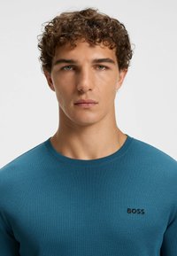 Teal long-sleeve shirt made of textured fabric with a round neck and black 'BOSS' logo on the chest. Simple design with a close fit.