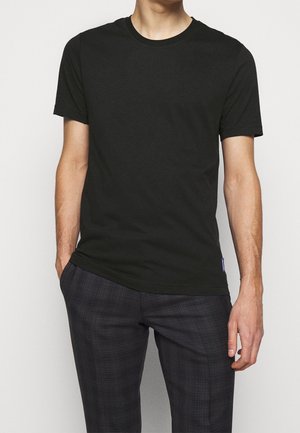 Mann trägt ein schlichtes schwarzes T-Shirt mit kurzen Ärmeln und dunkle karierte Hose, steht mit einer Hand in der Tasche vor einem schlichten Hintergrund.