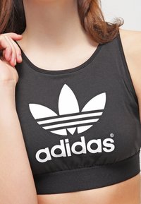 Top corto sportivo nero realizzato in un tessuto morbido, con un grande logo Adidas bianco e tre righe orizzontali sotto di esso.