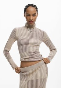 Top de cuello alto beige acanalado con rayas verticales en tonos más claros y oscuros, combinado con una falda texturizada a juego. Collar plateado con colgante en forma de corazón.