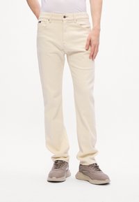 Personne portant un jean crème et des baskets beiges, se tenant contre un fond blanc uni, la main reposant sur la cuisse.