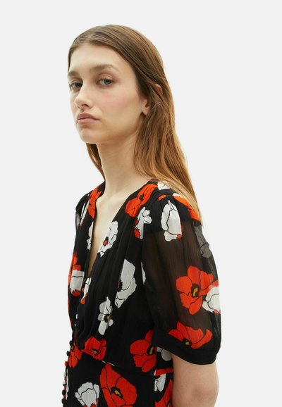 Junge Frau mit langen braunen Haaren, die ein schwarzes Kleid mit rotem und weißem Blumenmuster, durchsichtigen Puffärmeln und Knöpfen an der Vorderseite trägt.