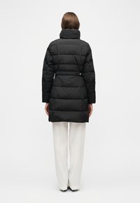 Άτομο που φοράει μακρύ μαύρο καπιτονέ παλτό με ψηλό γιακά, σε συνδυασμό με λευκό παντελόνι φαρδιάς γραμμής, στέκεται με την πλάτη προς τα εμπρός σε λευκό φόντο.