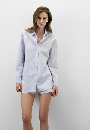 Kvinde stående op ad en ensfarvet væg iført en lyseblå stribet oversized skjorte med knapper og matchende shorts.