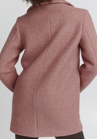 Femme portant un manteau en laine texturé rose poussiére, vue de dos, les mains posées sur les hanches, sur un fond uni.