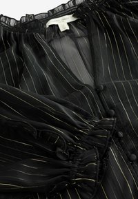 Blusa nera trasparente con strisce verticali dorate. Presenta un collo e polsini smock, bottoni decorativi e una texture di tessuto fluido.