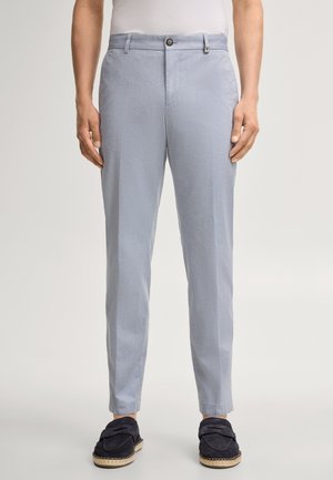 Mężczyzna w jasnoniebieskich dopasowanych spodniach i czarnych wsuwanych butach, stojący na jednolitym tle.