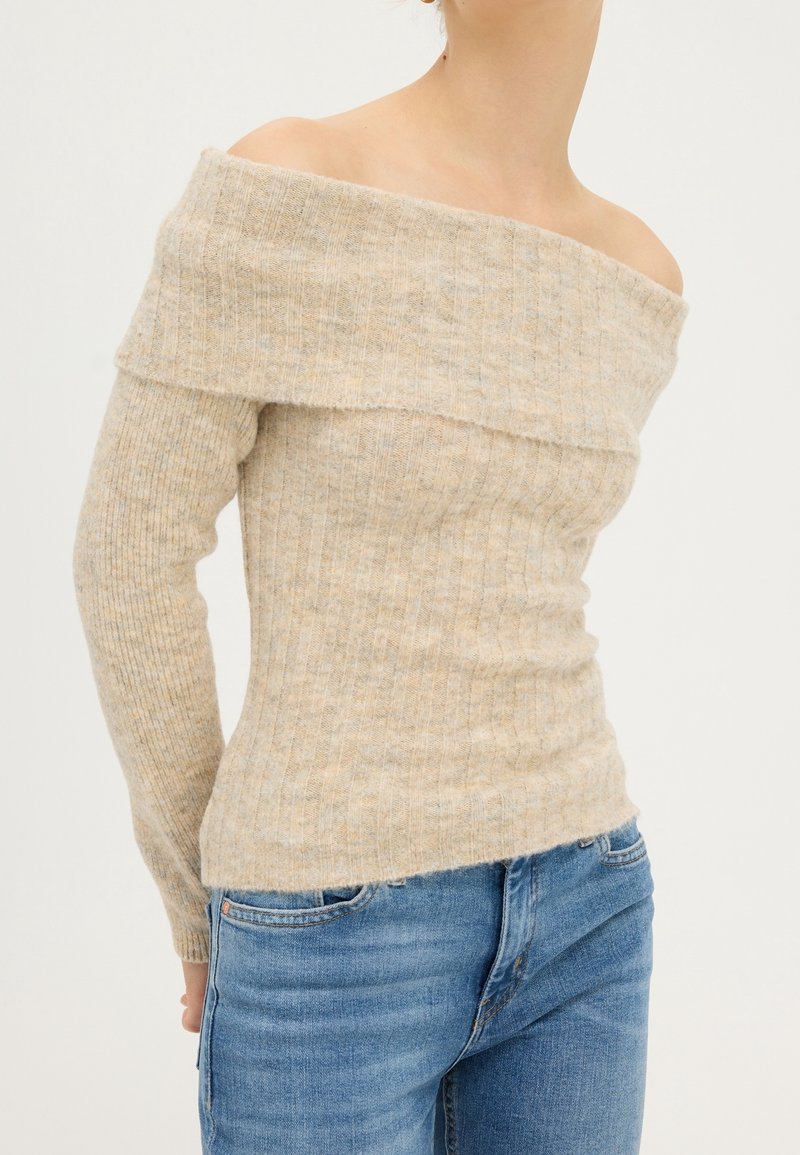 Pull beige côtelé à épaules dénudées porté par une personne, associé à un jean en denim bleu sur un fond uni.