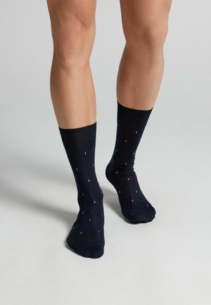 Beine mit schwarzer Wadenhoher Socke mit kleinem rot-weiß gepunktetem Muster, stehend auf einem hellgrauen Boden.