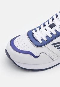 Biała tenisówka z niebieskim akcentem, chropowatą skórzaną cholewką, perforowanym czubkiem, białymi sznurowadłami oraz gumową podeszwą o zakrzywionym kształcie.