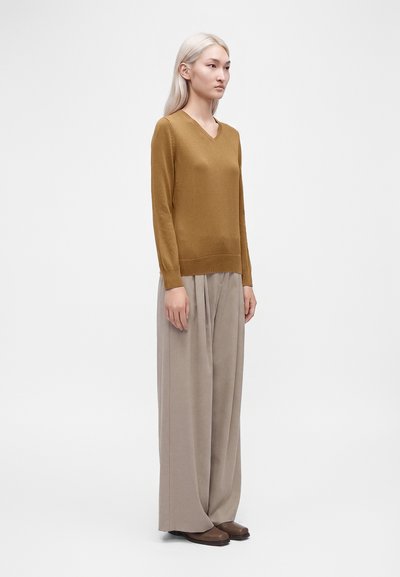 Femme aux longs cheveux clairs portant un pull moutarde à col en V, un pantalon large beige et des chaussures marron, debout sur un fond blanc uni.