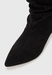 Botte de cheville en daim noir avec un bout pointu et une texture douce. Le design présente un tige ajustée sans quincaillerie ou motifs visibles.