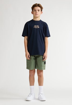 Camiseta de algodón azul marino con coordenadas topográficas en naranja, combinada con pantalones cortos cargo verdes y zapatillas blancas, con tela texturizada y bolsillos.