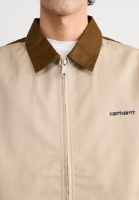 Beige ritsjack met een bruine kraag, gemaakt van gladde stof, met een klein geborduurd logo aan de linkerzijde.