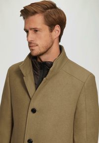 Man met lichtbruin haar, dragend een beige jas over een zwarte gewatteerde jack, kijkend naar links tegen een vlakke achtergrond.