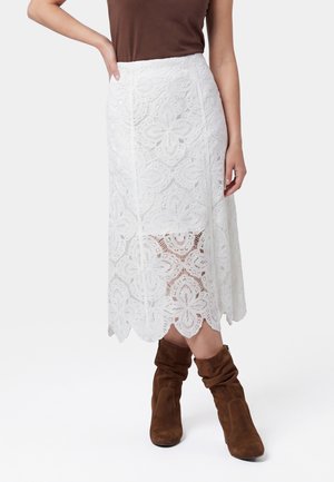 Frau trägt einen weißen Spitzen-Midirock mit Blumenmuster, braunes ärmelloses Top und braune lockere Stiefeletten vor schlichtem Hintergrund.