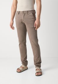 Beige Baumwoll-Chinos im Slim-Fit, mit seitlichen Taschen, einem Reißverschluss und einer dezenten, gewaschenen Textur, kombiniert mit hellbeigen Sandalen.