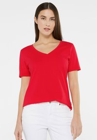 Red short-sleeve t-shirt with a V-neck design, made of smooth fabric, featuring a slightly relaxed fit. Worn with white jeans.