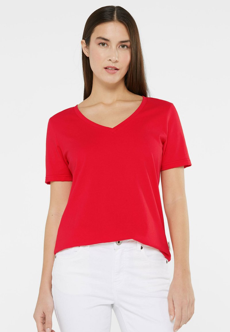 Red short-sleeve t-shirt with a V-neck design, made of smooth fabric, featuring a slightly relaxed fit. Worn with white jeans.