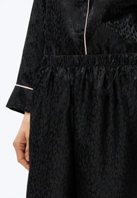 Ręka spoczywająca wzdłuż ciała, ubrana w czarny satynowy komplet piżamowy z delikatnym nadrukiem w panterkę i jasnoróżową lamówką na mankiecie i przednim szwie.